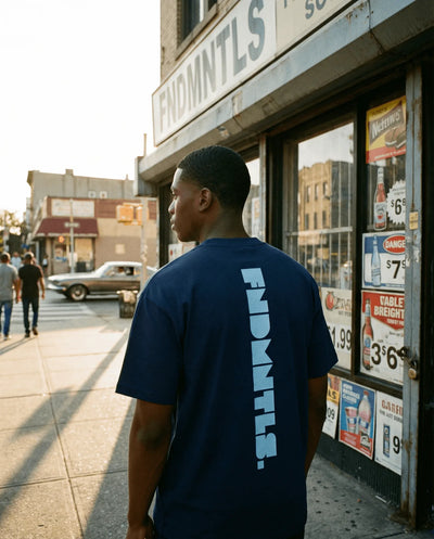 Man wearing a navy blue t-shirt with 'FNDMNTLS' branding standing on a city street.