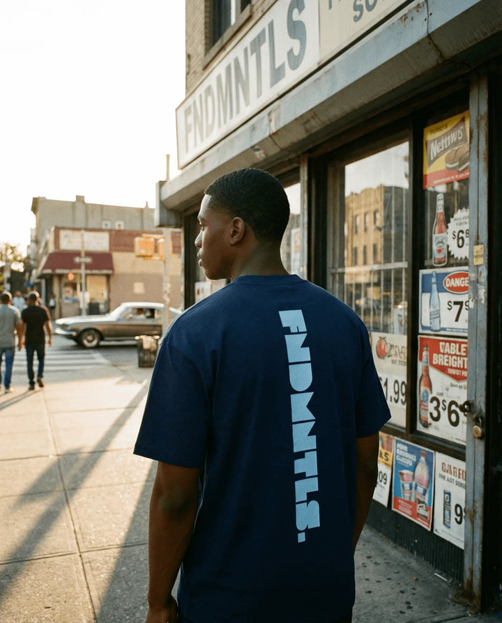 Man wearing a navy blue t-shirt with 'FNDMNTLS' branding standing on a city street.