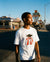 Person wearing a white t-shirt with a graphic design in front of a diner and motel sign.