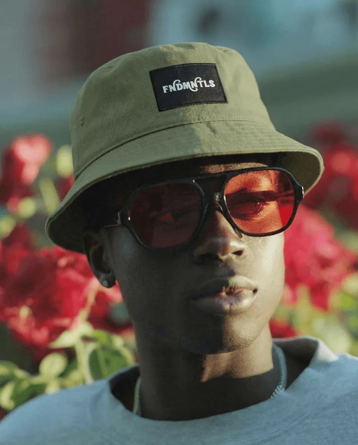 Person wearing a beige bucket hat with a brand logo and sunglasses, standing in front of red flowers.