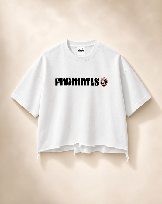 A white cropped t-shirt with a raw, frayed hem, featuring the text 'FNDMNTLS' in black across the chest alongside a small red and black graphic. The t-shirt is displayed flat against a warm beige background.