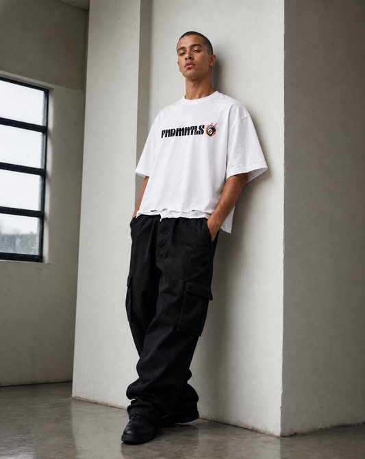 A man wearing a white t-shirt with a black and orange 'PHOENIXYS' graphic, paired with loose black cargo pants, leaning against a concrete pillar in a minimalist room.