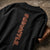 A black short-sleeve t-shirt laid flat on a textured concrete surface, featuring a vertical graphic print of the word 'STRENGTH' in a distressed copper-brown block font.