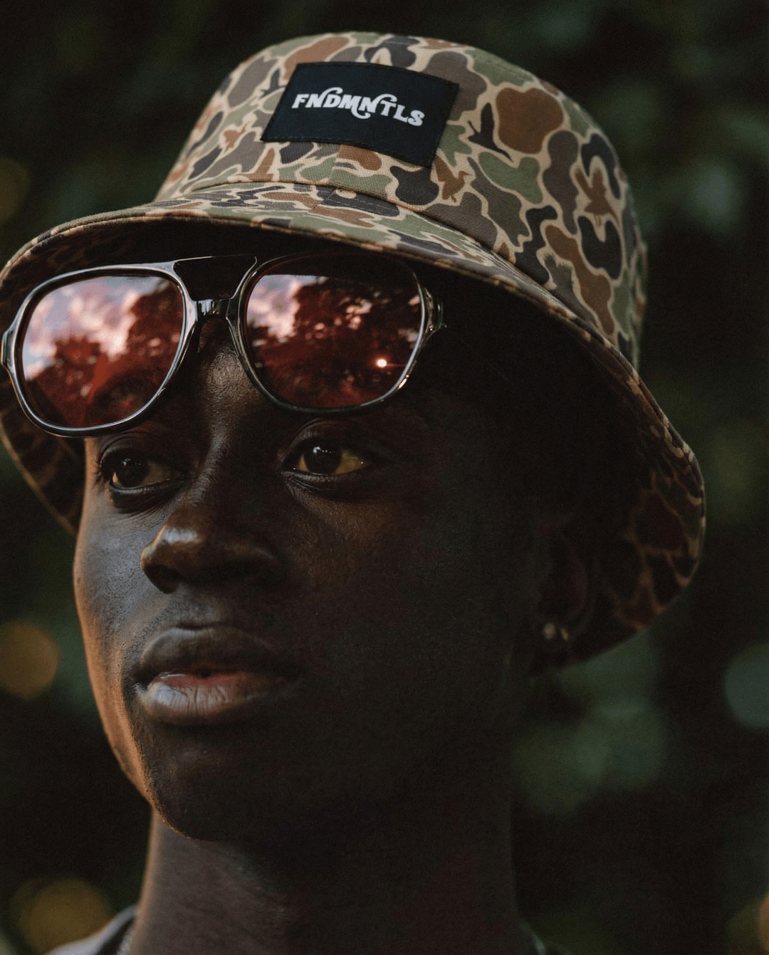 Person wearing a camouflage bucket hat with a brand logo and reflective sunglasses, against a blurred natural background.