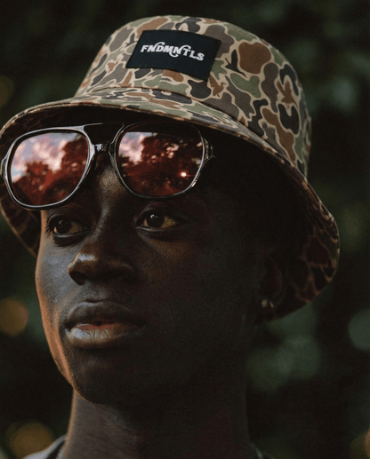 Person wearing a camouflage bucket hat with a brand logo and reflective sunglasses, against a blurred natural background.
