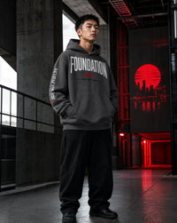 A man wearing a dark gray hoodie with 'FOUNDATION' printed in white across the chest and black pants, standing in a modern industrial interior with concrete pillars and red ambient lighting.