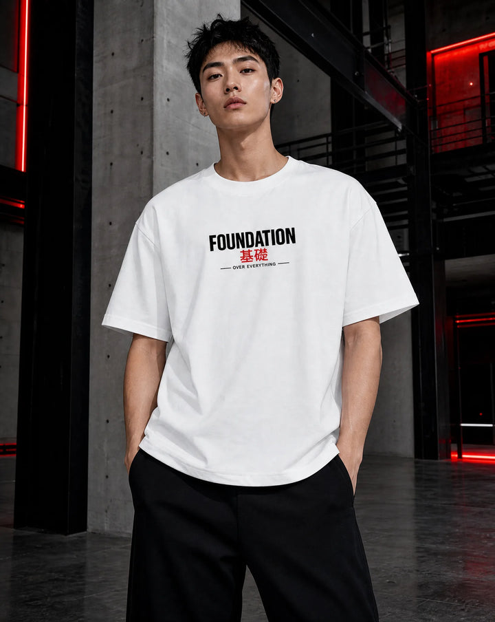 A man wearing a white short-sleeve t-shirt with 'FOUNDATION' printed in black and red on the chest, paired with black pants, standing in a modern industrial interior with red ambient lighting.