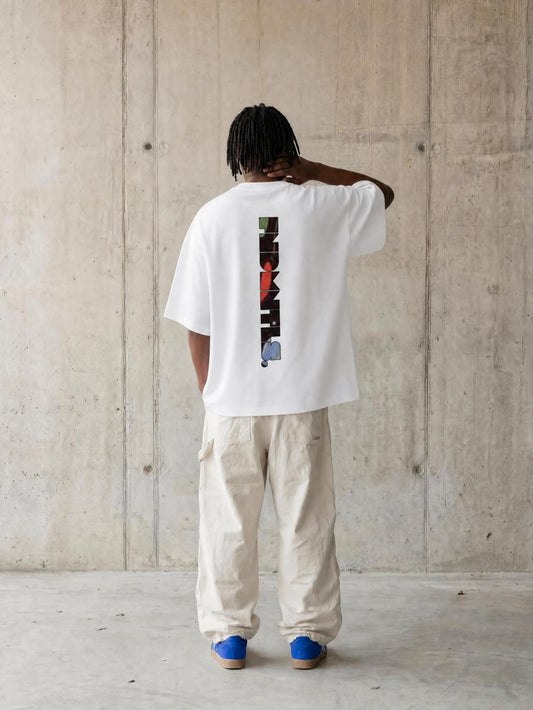 A person standing with their back to the camera wearing a white oversized t-shirt with a vertical graphic print on the back, paired with beige cargo pants and blue shoes, set against a concrete wall.