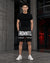 A man stands in a modern industrial setting wearing a black t-shirt and black shorts with bold white text reading 'FNDMNTLS' and white sneakers.