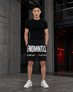 A man stands in a modern industrial setting wearing a black t-shirt and black shorts with bold white text reading 'FNDMNTLS' and white sneakers.