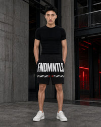 A man stands in a modern industrial setting wearing a black t-shirt and black shorts with bold white text reading 'FNDMNTLS' and white sneakers.