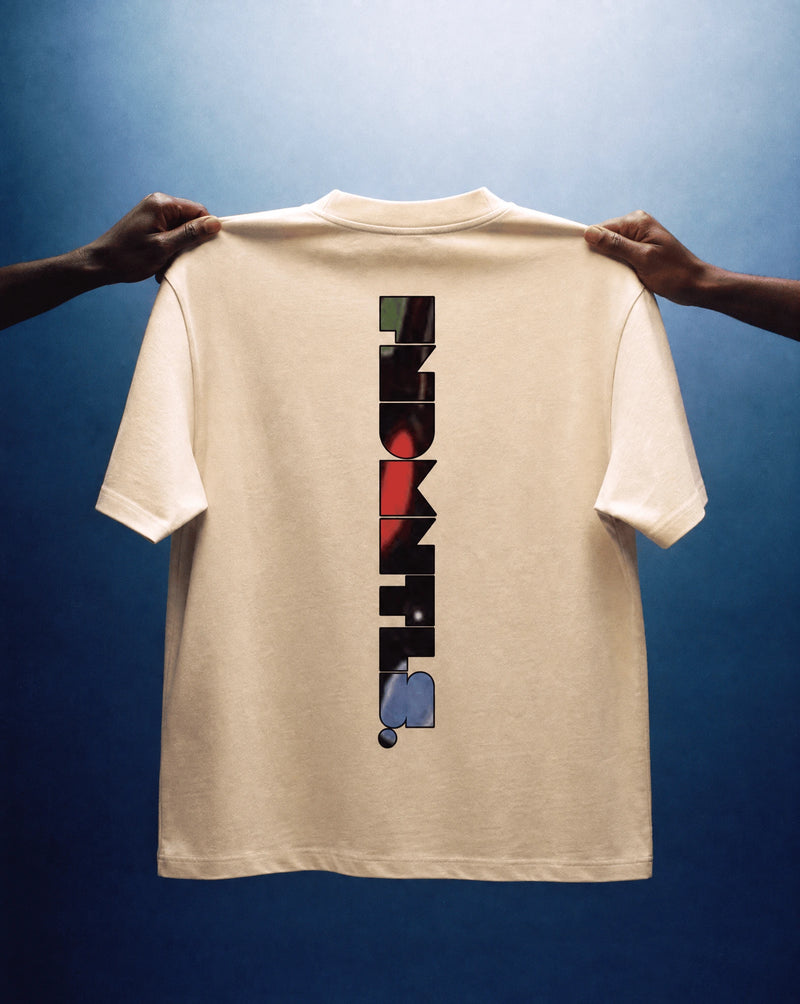 A cream-colored t-shirt is held up by two hands against a dark blue gradient background. The back of the shirt features a vertical graphic spelling out 'HOKIT' in black blocks with small accent shapes in red, green, and blue.
