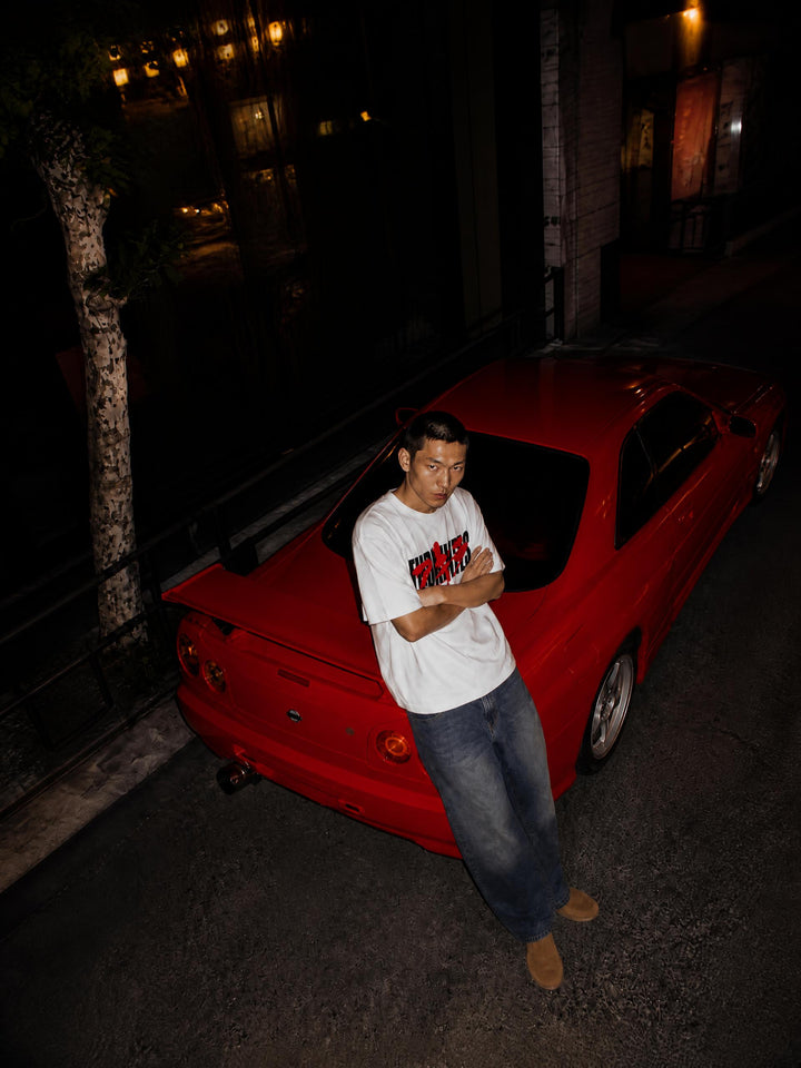 A man wearing a white graphic t-shirt and blue jeans stands next to a red sports car at night on a street.