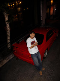A man wearing a white graphic t-shirt and blue jeans stands next to a red sports car at night on a street.