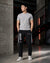 A man stands in a modern industrial loft wearing a light gray crewneck t-shirt, black track pants with white side stripes and Chinese characters, and white sneakers.