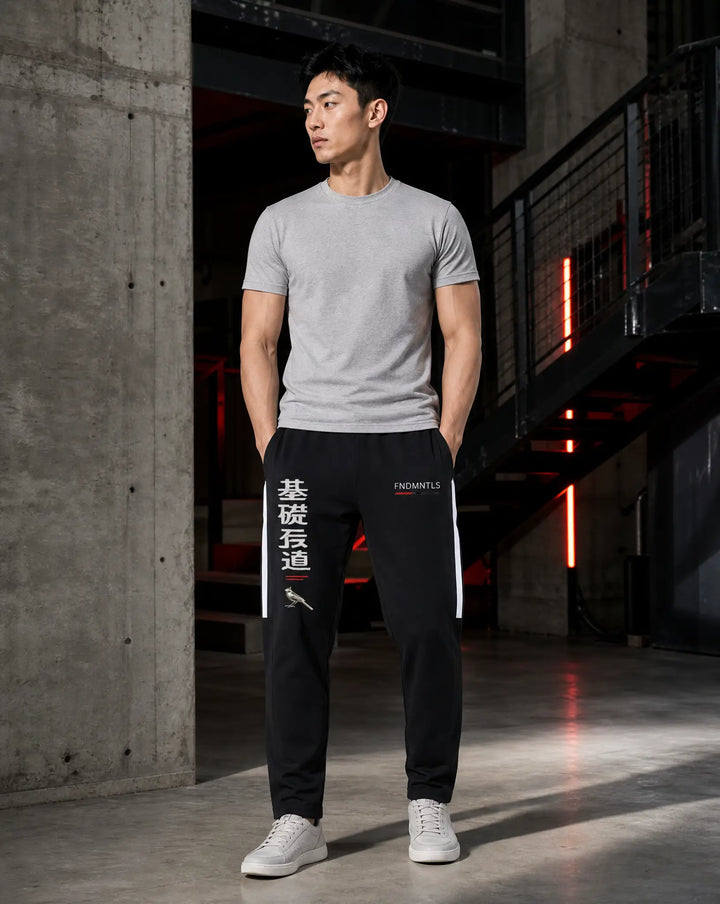 A man stands in a modern industrial loft wearing a light gray crewneck t-shirt, black track pants with white side stripes and Chinese characters, and white sneakers.