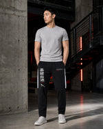 A man stands in a modern industrial loft wearing a light gray crewneck t-shirt, black track pants with white side stripes and Chinese characters, and white sneakers.