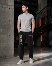 A man stands in a modern industrial loft wearing a light gray crewneck t-shirt, black track pants with white side stripes and Chinese characters, and white sneakers.