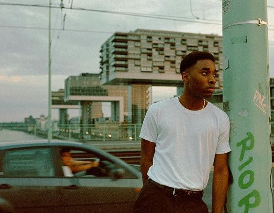Man in a white t-shirt standing next to a car with a cityscape in the background