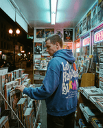 Let Happiness Bloom Vintage Wash Frayed Fleece Hoodie in blue worn by man browsing magazines in a retro store. Casual streetwear with floral graphic back.