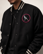 A close-up of a person wearing a black varsity-style jacket with striped ribbed collar and cuffs, silver snap buttons, and a circular embroidered bird patch in pink and gray on the left chest.