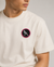 A close-up of a person wearing a cream-colored crewneck t-shirt with a circular chest patch featuring a bird design.
