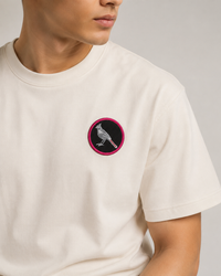 A close-up of a person wearing a cream-colored crewneck t-shirt with a circular chest patch featuring a bird design.