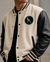 A close-up of a person wearing a cream and black varsity-style jacket with a circular bird patch on the chest, over a black shirt.