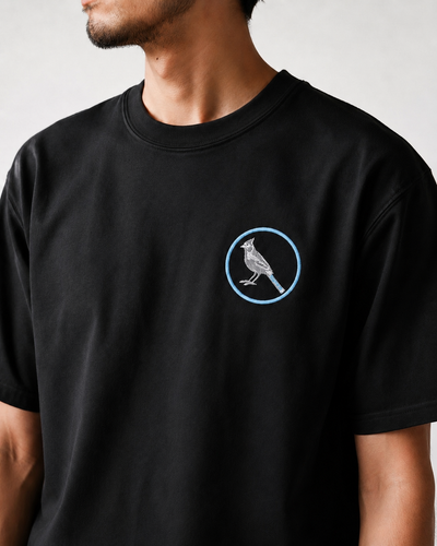 A man wearing a black crewneck t-shirt with a small graphic on the left chest showing a white bird inside a light blue circle, set against a plain light gray background.