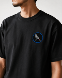 A man wearing a black crewneck t-shirt with a small blue and white bird graphic on the left chest, set against a plain light gray background.