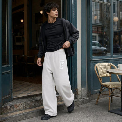 A man stands on the sidewalk outside a storefront wearing loose white sweatpants with a large white letter 'F' on the thigh, a black t-shirt, a black zip-up jacket, and black shoes. A woven chair and cafe table are visible to the right.