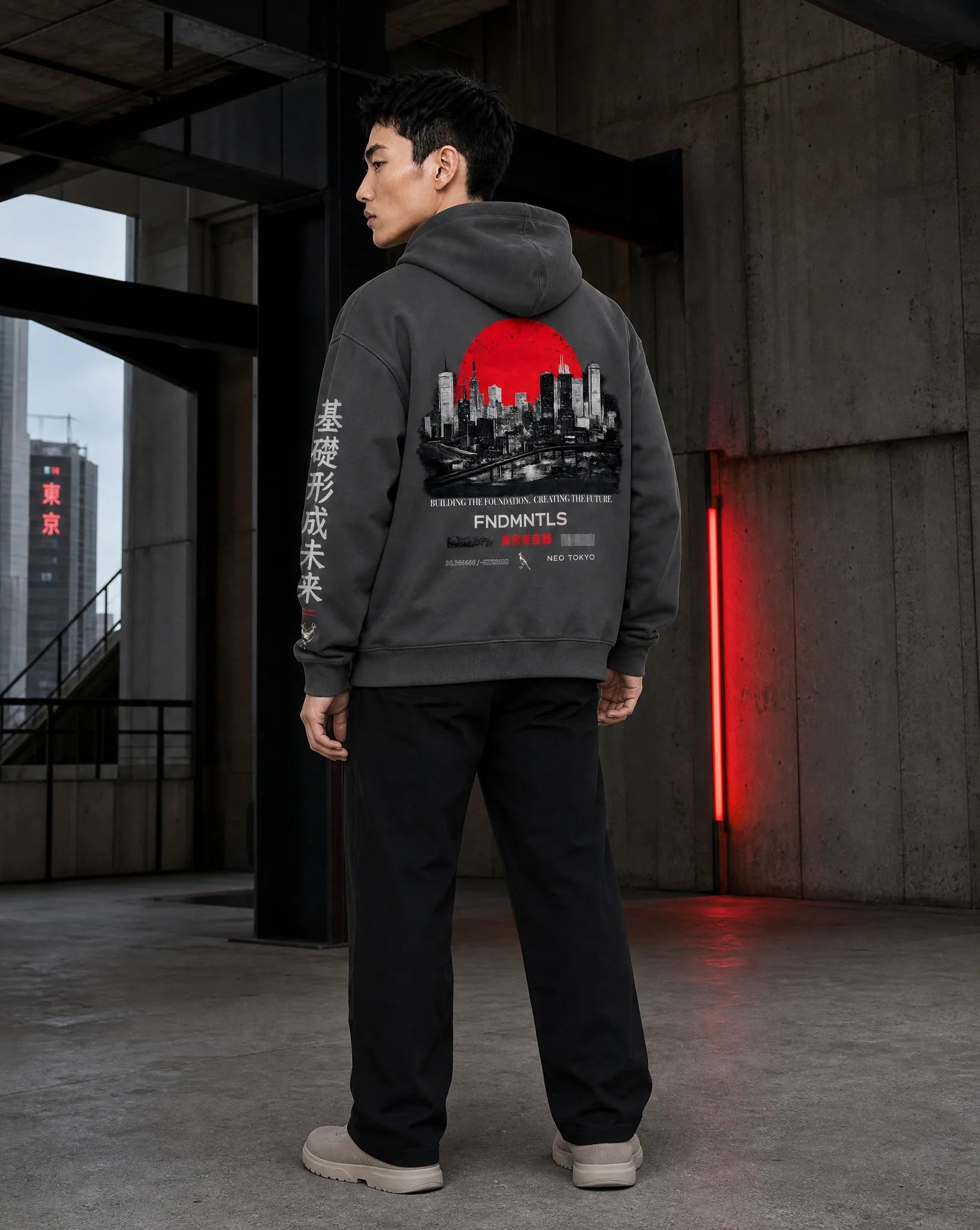 A man wearing a dark gray hoodie with a large red and black city graphic on the back, standing in a concrete industrial interior.