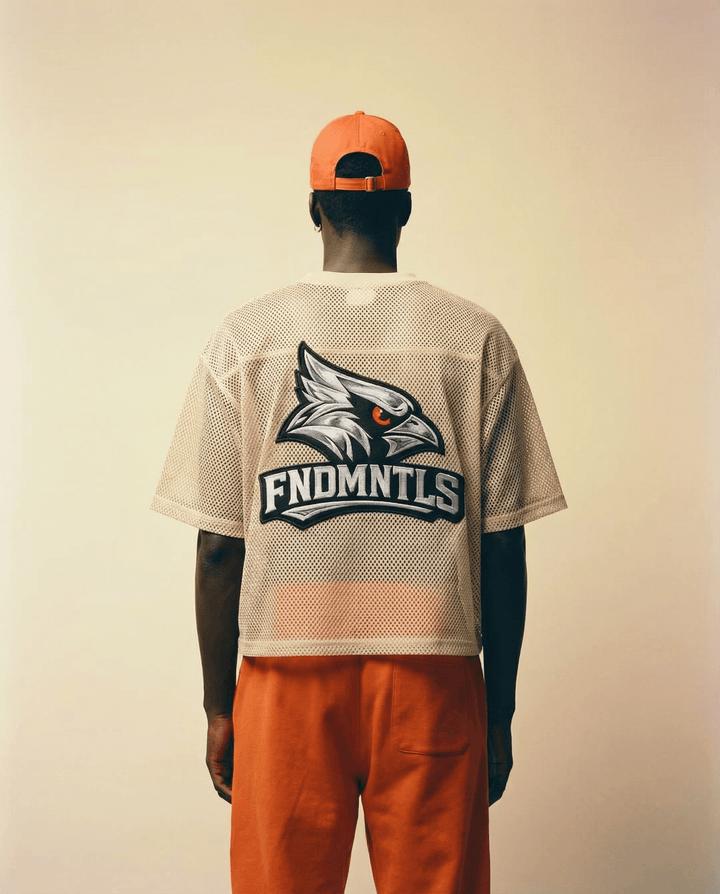 Person wearing a beige sports jersey with a logo and text, orange pants, and an orange cap on a plain background