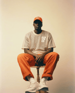 Person wearing an orange cap, beige t-shirt with a logo, and orange pants sitting on a stool.