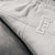 Close-up of ribbed light gray sweatpants with a large white and gray embroidered monogram logo on the thigh, resting on a dark textured surface.