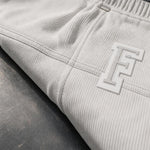 Close-up of ribbed light gray sweatpants with a large white and gray embroidered monogram logo on the thigh, resting on a dark textured surface.