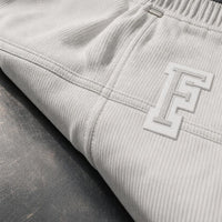 Close-up of ribbed light gray sweatpants with a large white and gray embroidered monogram logo on the thigh, resting on a dark textured surface.