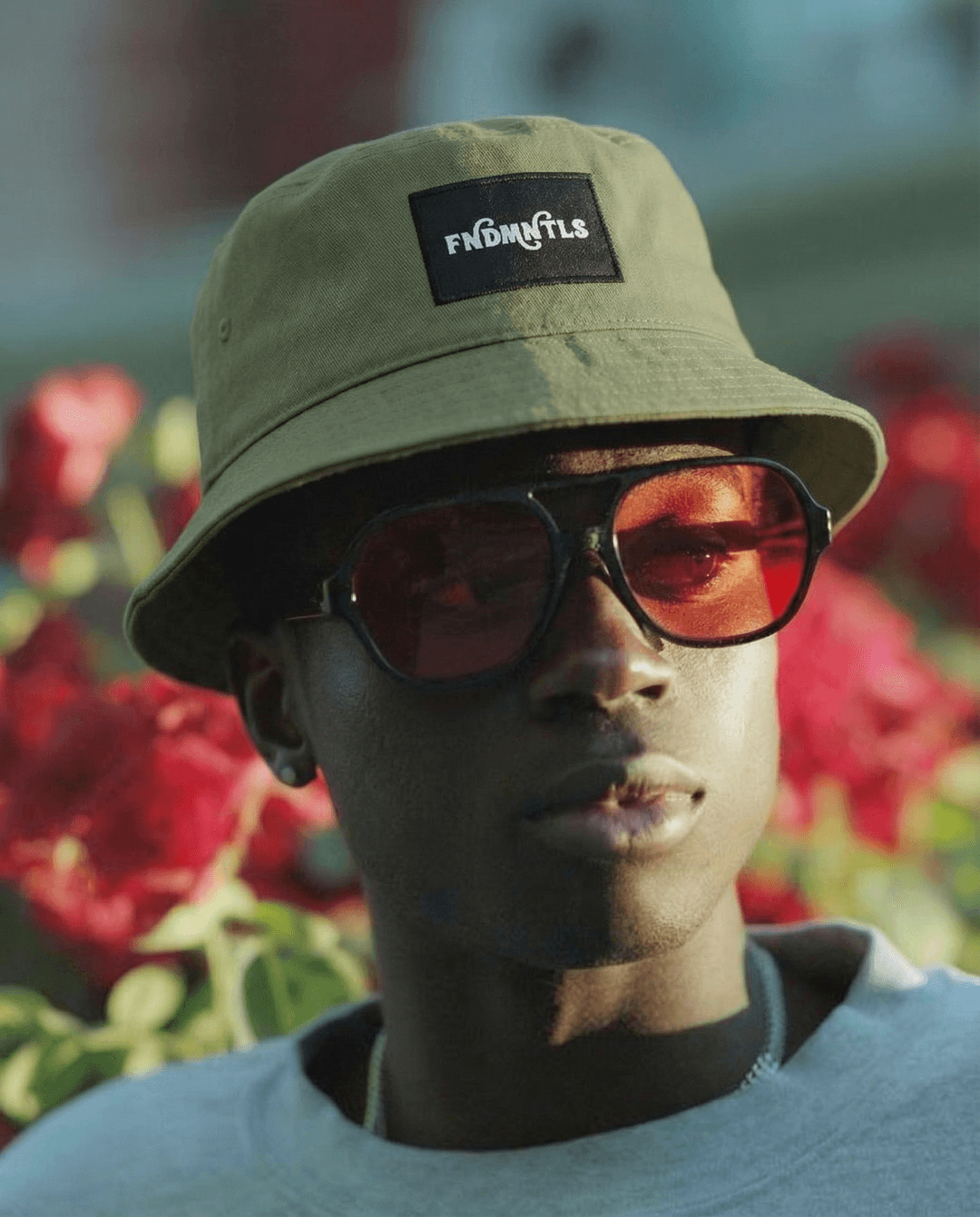 Person wearing a beige bucket hat with a brand logo and sunglasses, standing in front of red flowers.