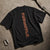 A black short-sleeve t-shirt laid flat on a textured concrete surface, showing the back with a vertical graphic print in a distressed orange-brown color. A silver carabiner and a strap are placed nearby as props.