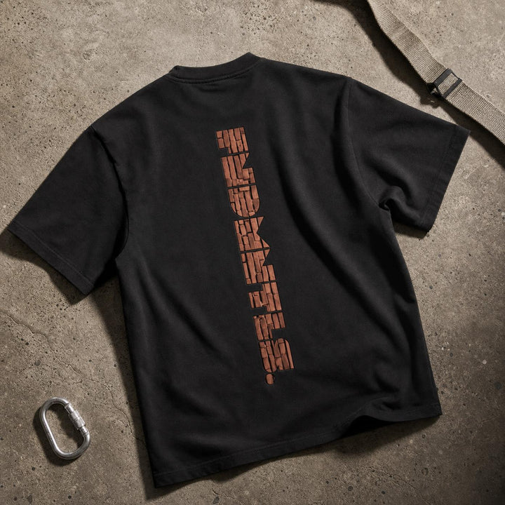 A black short-sleeve t-shirt laid flat on a textured concrete surface, showing the back with a vertical graphic print in a distressed orange-brown color. A silver carabiner and a strap are placed nearby as props.