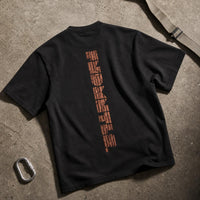 A black short-sleeve t-shirt laid flat on a textured concrete surface, showing the back with a vertical graphic print in a distressed orange-brown color. A silver carabiner and a strap are placed nearby as props.