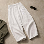 White textured knit pants with a large white outlined letter 'F' on the upper left leg, styled flat-lay on a beige stone surface with black sunglasses and an olive green garment partially visible.