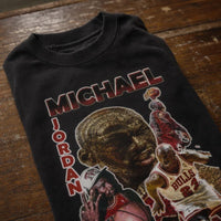 A black graphic t-shirt featuring a collage of Michael Jordan in red, white, and gold, with 'MICHAEL JORDAN' printed in red and white text, laid flat on a rustic wooden surface.