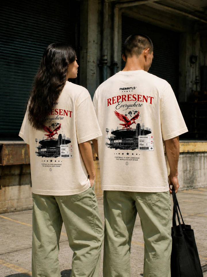 Two models standing side-by-side from behind wearing matching cream-colored t-shirts with a large red and black graphic print on the back, paired with light green cargo pants in an urban alleyway.
