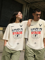 A man and a woman wearing matching off-white graphic t-shirts with 'WORLD STAGE' text and a globe design, paired with olive green cargo pants, standing in an urban alleyway.