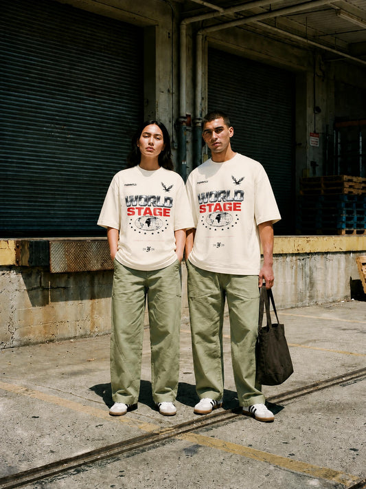 A man and a woman stand outdoors in front of a warehouse-style building, wearing matching off-white graphic T-shirts with a red and black 'WORLD STAGE' print, paired with light olive pants and white sneakers.