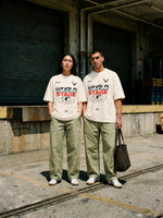 A man and a woman stand outdoors in front of a warehouse-style building, wearing matching off-white graphic T-shirts with a red and black 'WORLD STAGE' print, paired with light olive pants and white sneakers.