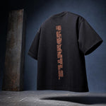 A black oversized short-sleeve t-shirt shown from the back, featuring a vertical metallic copper-orange graphic print on the center back. The shirt is displayed against a dark blue-gray studio background with a textured metal column prop on the left.