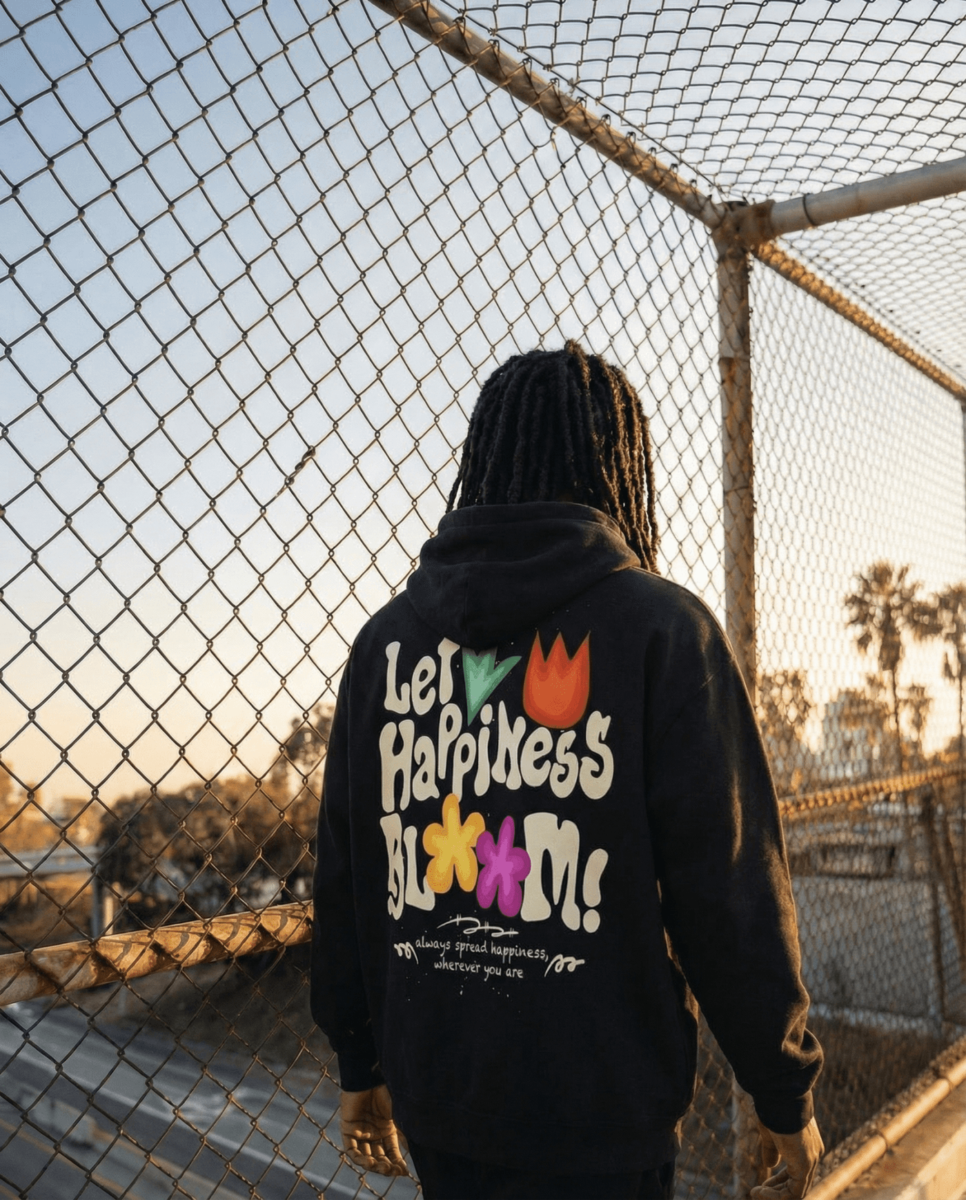 Let Happiness Bloom Vintage Wash Frayed Fleece Hoodie in black worn outdoors near a fence. Oversized cotton hoodie with bold floral back print.