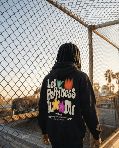 Let Happiness Bloom Vintage Wash Frayed Fleece Hoodie in black worn outdoors near a fence. Oversized cotton hoodie with bold floral back print.